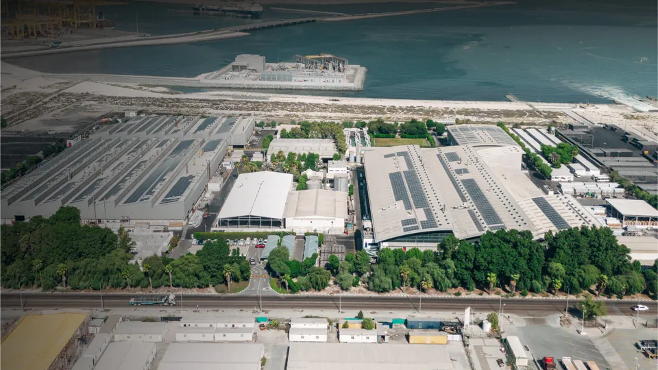 Aerial view of the Gulf Extrusion plant: long factory halls with solar panels on the roofs, palm trees along the road and the sea beyond.