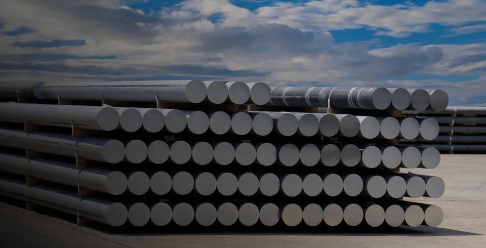 Stacks of round aluminium billets in a yard under a cloudy sky.